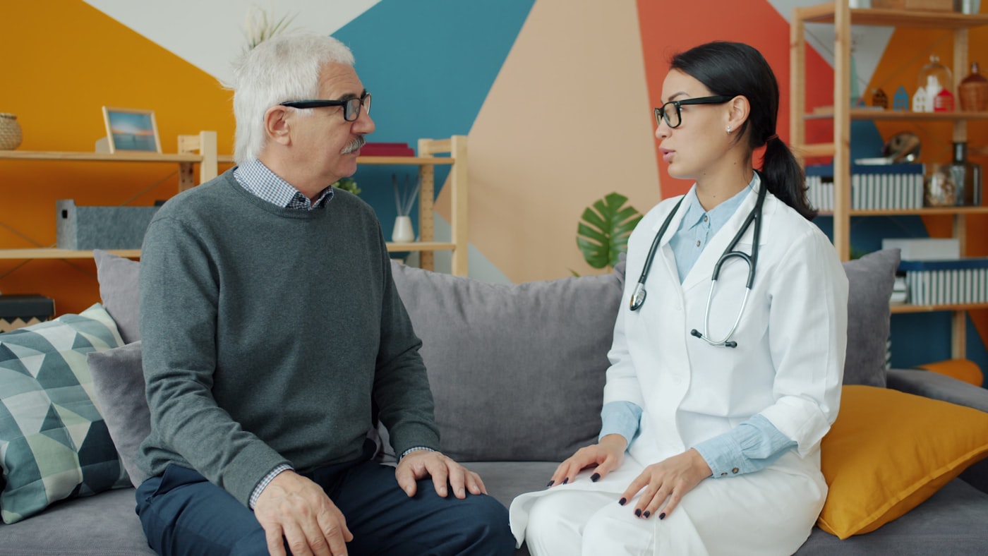A pharmacist in a relaxed conversation with an older adult on a sofa.